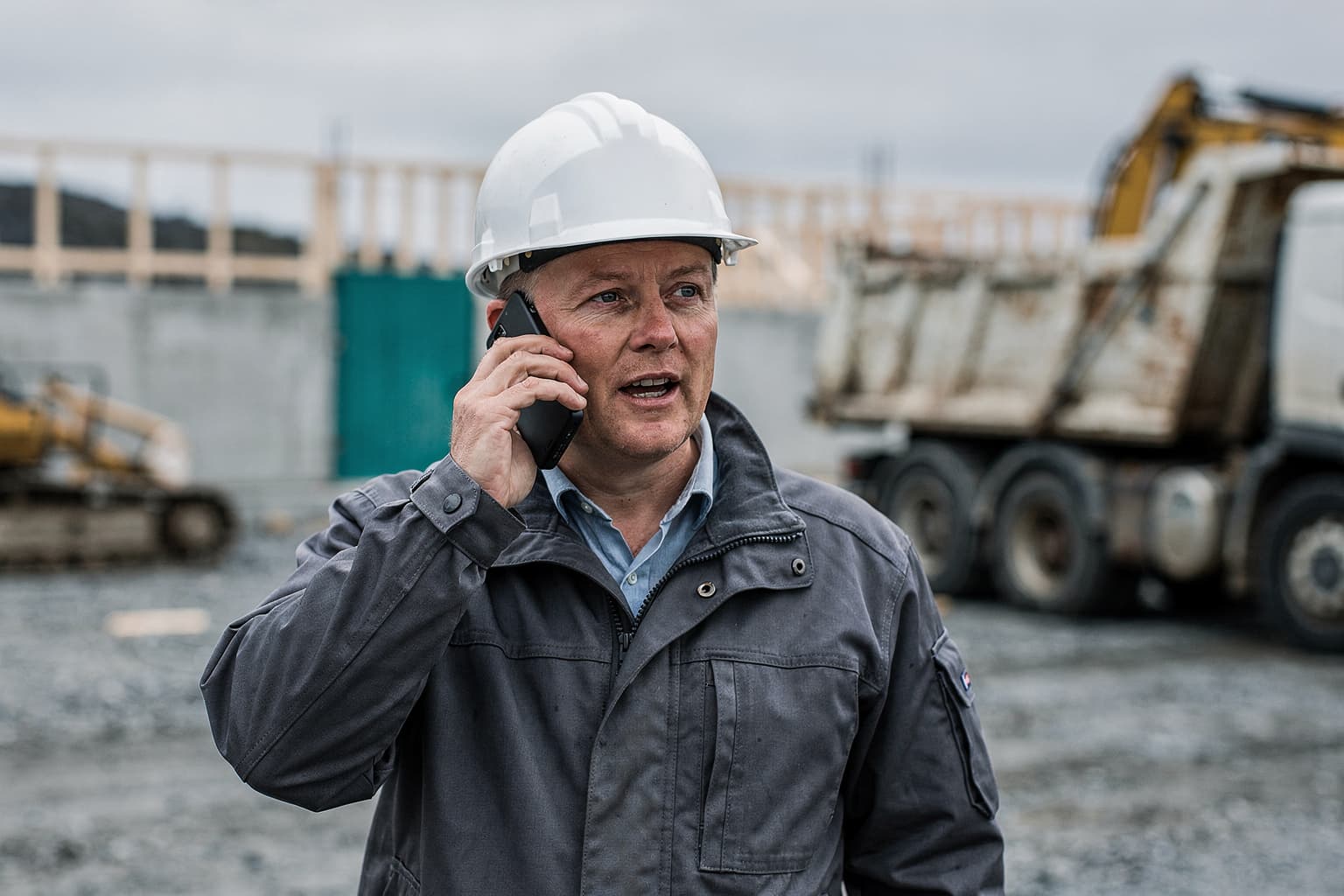 Byggeleder i telefonen på en byggeplass, med gravemaskin bak